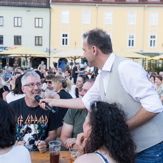 Ein Mann mit Mikrofon spricht zu einem Publikum im Freien. Das Publikum sitzt an Tischen. Im Hintergrund sind gelbe Sonnenschirme und ein gelbes Gebäude zu sehen.