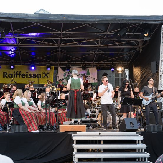 Eine Band tritt auf der Bühne auf, mit einer Sängerin in traditioneller Kleidung, einem Mann mit Sonnenbrille, der singt, und einem Gitarristen. Dahinter spielt eine Gruppe von Musikern Instrumente.