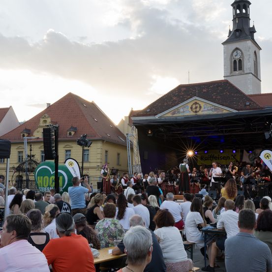 Eine Musikband spielt auf einer Bühne vor einem großen Publikum. Hinter der Bühne befindet sich ein historisches Gebäude mit einem Glockenturm. Die Menschen sitzen auf Bänken und Tischen und sehen sich die Aufführung an.