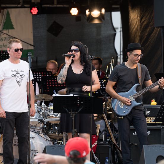 Eine Band tritt auf der Bühne auf, mit einem Sänger vorne, einem Gitarristen rechts und einem Schlagzeuger links. Das Publikum ist im Hintergrund sichtbar.