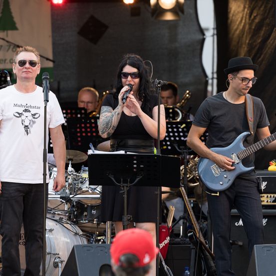 Eine Band tritt auf einer Bühne auf, mit einer Sängerin am Mikrofon, einem Gitarristen und einem Schlagzeuger. Im Hintergrund befinden sich Musikinstrumente und ein Publikum.