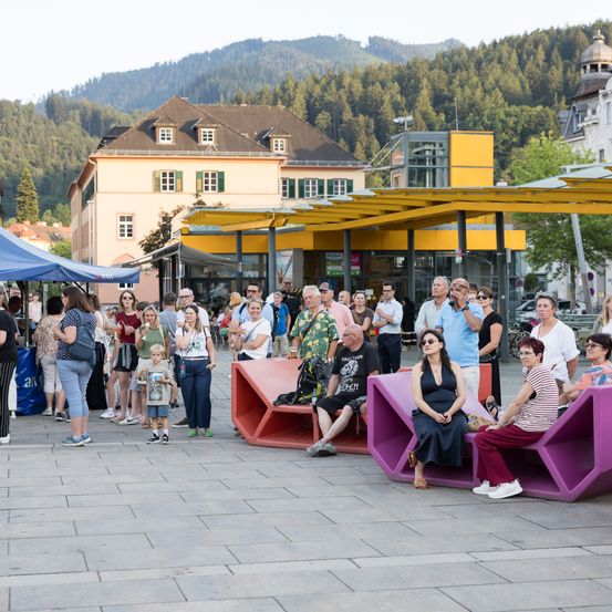 Eine Gruppe von Menschen steht und sitzt auf Bänken auf einem Stadtplatz, mit Bergen und Gebäuden im Hintergrund.