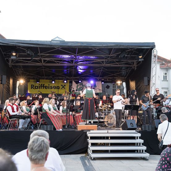 Eine Gruppe von Musikern tritt auf einer Bühne unter einem Zelt mit einem Banner mit dem Schriftzug Raiffeisen auf. Das Publikum sitzt davor.