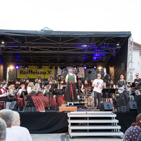 Eine Bühnenaufführung mit einer vielfältigen Band. Musiker in traditioneller Kleidung, moderne Instrumente und ein Banner mit der Aufschrift 'Raiffeisen'. Das Publikum schaut zu.