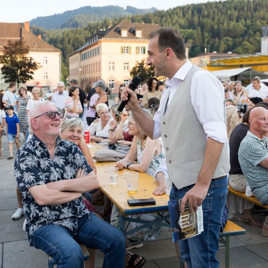 Ein Mann spricht in ein Mikrofon bei einer überfüllten Veranstaltung im Freien. Mehrere Personen sitzen und stehen herum, genießen Getränke und Gespräche. Im Hintergrund sind Berge sichtbar.