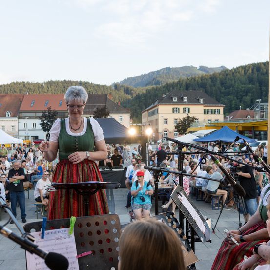Eine Frau in traditioneller Kleidung dirigiert eine Musikaufführung. Viele Menschen sitzen und stehen hinter ihr und genießen die Musik. Ein Gebirgshintergrund ist in der Ferne sichtbar.