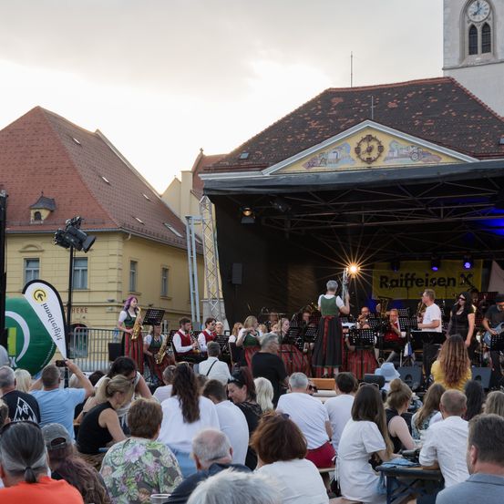 Eine Musikband tritt auf der Bühne auf, während eine große Menschenmenge zuschaut. Die Bühne hat ein Sponsorenbanner und Lichter. Das Publikum ist vielfältig, sitzt und steht. Der Hintergrund besteht aus Gebäuden und einem bewölkten Himmel.