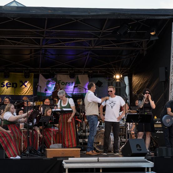 Eine Band tritt auf der Bühne mit Bannern im Hintergrund auf. Zwei Männer interagieren auf der Bühne, einer hält ein Mikrofon. Verschiedene Musiker und Sänger sind anwesend. Musikinstrumente und Lautsprecher sind sichtbar.