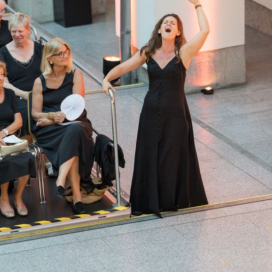 Eine Frau singt auf einer Bühne, während mehrere Frauen im Publikum sitzen, eine hält einen Fächer. Sie tragen alle schwarze Kleider.
