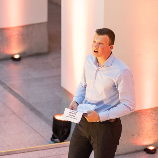 Ein Mann steht auf einem Betonboden und hält ein Stück Papier mit Text in der Hand. Er spricht in ein Mikrofon, möglicherweise bei einer Veranstaltung. Auf dem Boden befinden sich zwei kleine Lichter.