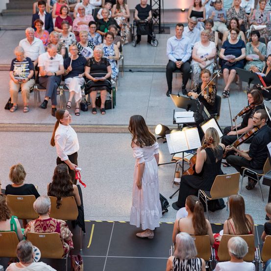 Ein Publikum älterer Erwachsener sieht ein klassisches Musikkonzert. Die auftretenden Musiker sind Geiger, Cellisten und ein Pianist. Das Publikum sitzt auf Stühlen, manche halten Programme in den Händen.
