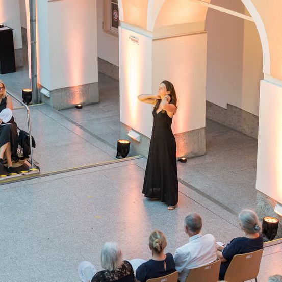 Eine Frau in einem schwarzen Kleid steht auf einer Bühne und tritt für ein kleines Publikum in einer modernen Indoor-Veranstaltung auf.