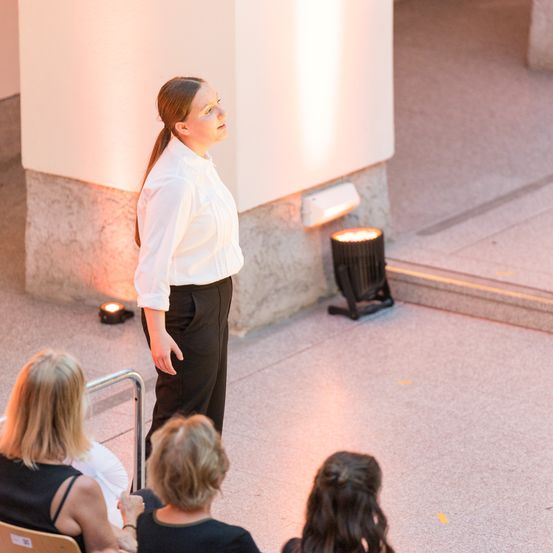 Eine Frau in einer weißen Bluse und schwarzen Hosen steht mit erhobenem Blick. Mehrere Frauen sitzen unter ihr und schauen zu. Der Bereich ist gut beleuchtet mit Scheinwerfern an den Wänden.