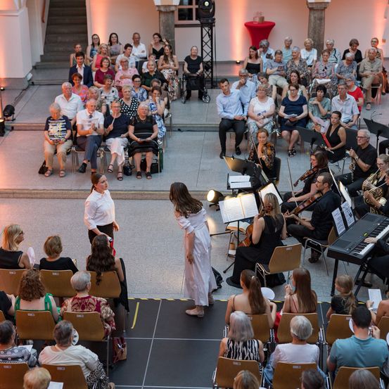 Ein Orchester spielt auf der Bühne vor einem sitzenden Publikum. Eine Frau in einem weißen Kleid steht zwischen den Musikern und dem Publikum, möglicherweise spricht oder stellt sie sie vor. Das Publikum beobachtet aufmerksam die Aufführung.