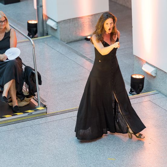 Eine Frau in einem schwarzen Kleid zeigt auf die Seite und steht auf einem gefliesten Boden, mit einer anderen Frau im Sitzen hinter ihr.