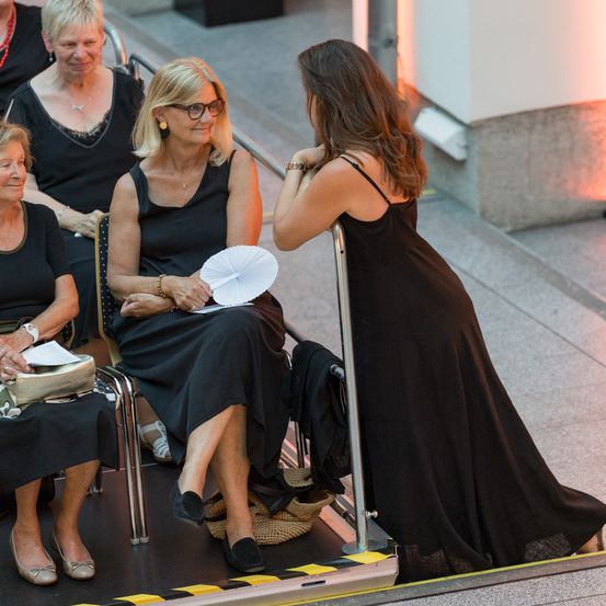 Fünf Frauen sitzen auf einer Bühne. Eine Frau hält ein Stück Papier, während eine andere Frau auf sie zukommt.