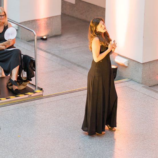 Eine Frau in einem schwarzen Kleid singt auf einer Bühne, mit einer sitzenden Frau im Hintergrund.