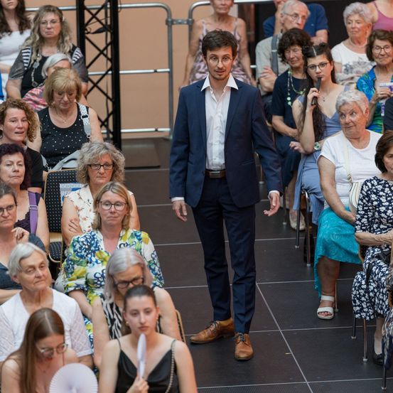 Ein Mann in einem Anzug geht an einem Publikum sitzender Frauen vorbei.