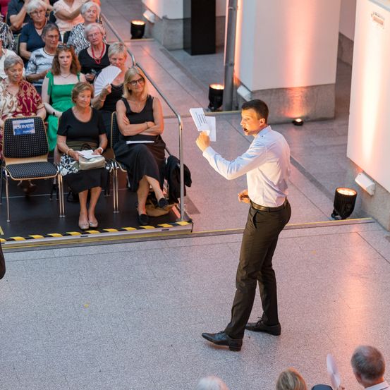 Ein Mann steht auf der Bühne und spricht zu einem Publikum sitzender Frauen. Er hält ein Papier in der Hand und scheint eine Rede zu halten.