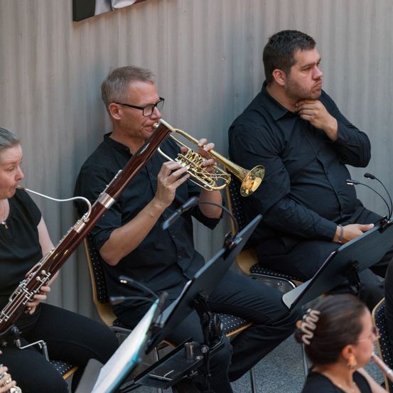 Eine Gruppe von Musikern in schwarzen Hemden spielt Instrumente. Einer spielt die Trompete, und andere spielen Fagott und Flöte. Sie sitzen auf Stühlen mit Notenständern.