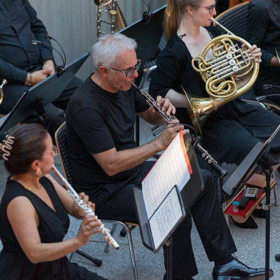 Eine Band tritt auf, wobei ein Mann die Klarinette, eine Frau die Flöte und eine weitere Frau die französische Horn spielt. Sie sitzen auf Stühlen mit Notenständern vor ihnen.