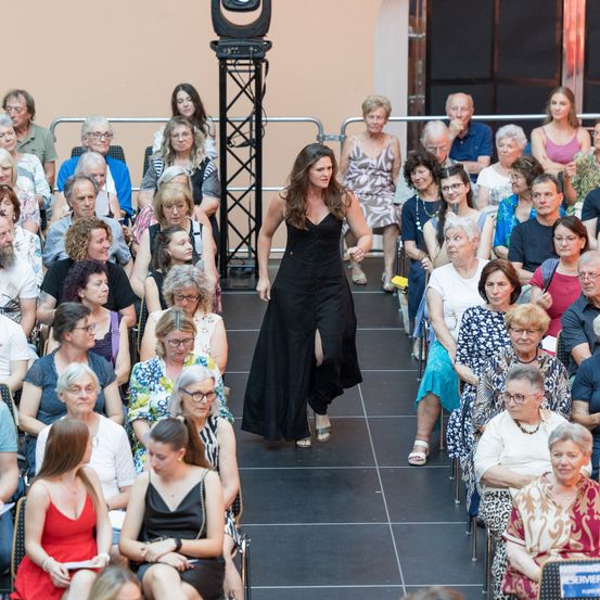 Eine Frau in einem schwarzen Kleid geht auf die Bühne, vor ihr sitzt das Publikum. Das Publikum besteht aus verschiedenen Personen, einige tragen Brillen, andere sind lässig gekleidet. Ein Scheinwerfer befindet sich hinter der Bühne.