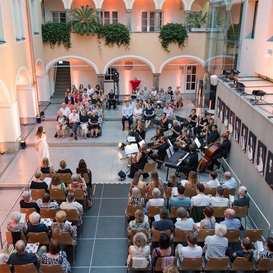 Ein klassisches Musikensemble spielt in einem Innenhof, mit einem Solisten auf der linken Seite. Das Publikum, meist sitzend, hört aufmerksam zu. Der Raum ist mit Rundbogenfenstern und Topfpflanzen ausgestattet.