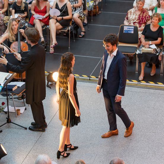 Ein Mann in einem Anzug geht an einer Frau in einem grünen Kleid auf der Bühne vorbei. Hinter ihnen spielt ein Mann Cello und dirigiert ein kleines Orchester. Zuschauer sitzen im Hintergrund.