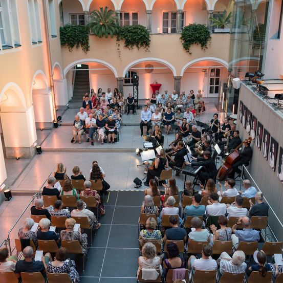 Ein Konzertsaal mit einem großen Publikum in Reihen. Musiker spielen auf der Bühne verschiedene Instrumente. Der Saal hat gewölbte Öffnungen und Topfpflanzen.