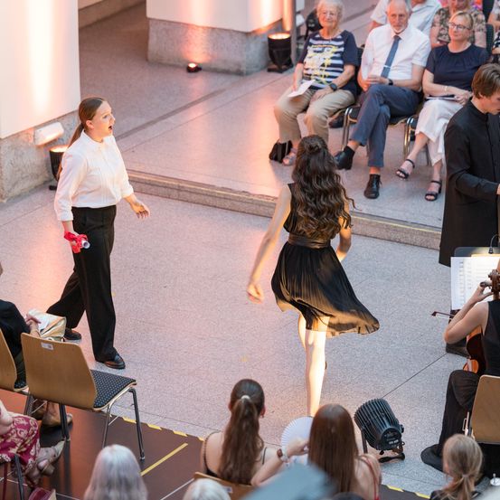 Eine Frau in einem weißen Hemd singt, während eine andere Frau in einem schwarzen Kleid vor einem sitzenden Publikum tanzt. Eine Violinistin und eine Cellistin spielen im Hintergrund.