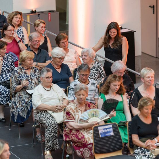 Eine Gruppe älterer Frauen sitzt in Stühlen in einem Theater, einige tragen Brillen, Uhren und Halsketten. Eine Frau in einem schwarzen Kleid steht in der Mitte und hält einen Fächer. Eine andere Frau sitzt mit einem Buch und einer Handtasche.
