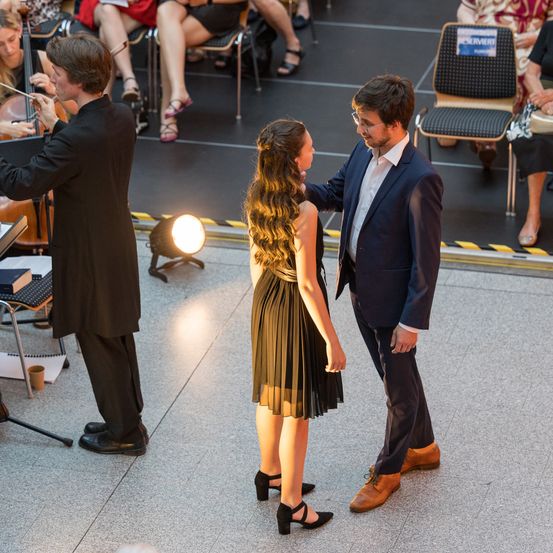 Ein Mann und eine Frau stehen auf einer Bühne vor einem Cellospieler. Der Mann hält die Hand der Frau und schaut sie an. Hinter ihnen sitzen Leute auf Stühlen und schauen sich die Aufführung an.