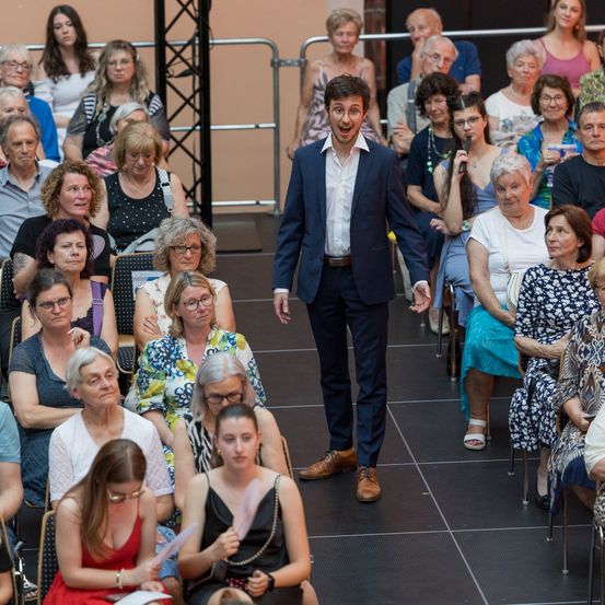 Ein Mann in einem Anzug geht über eine Bühne und lächelt das Publikum an. Dahinter sitzen mehrere ältere Frauen und Männer in Stühlen, einige tragen Brillen und Halsketten.