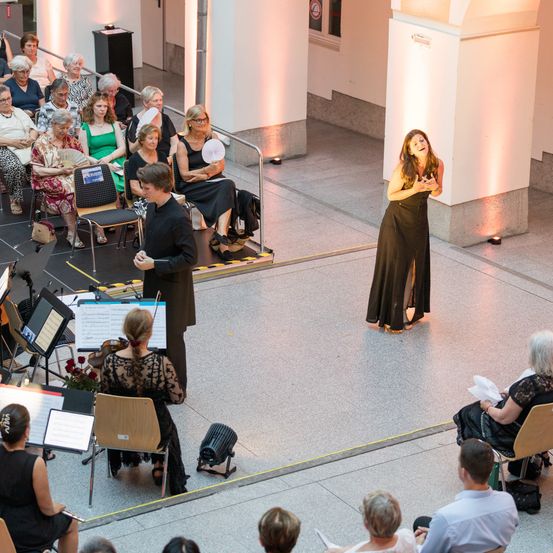 Eine Frau singt auf der Bühne, während eine Geigerin vor einem sitzenden Publikum spielt. Ein Mann steht neben ihr und hält einen Notenständer.