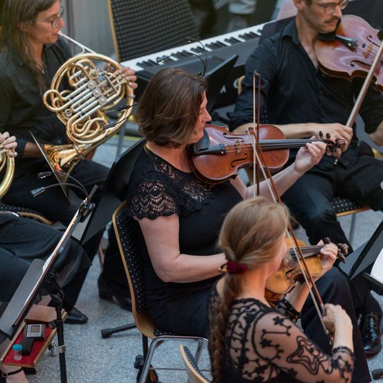 Eine Gruppe von Musikern in Schwarz spielt im Innenbereich. Eine Frau spielt die Violine, während ein Mann die französische Horn spielt. Ein Pianist und ein Violinist sitzen hinter ihnen. Der Musiker auf der linken Seite spielt die Trompete.