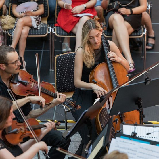 Eine Frau spielt Cello auf einem Stuhl, während zwei Männer vor ihr Violinen spielen. Dahinter hält eine Frau ein Notenständer mit Papieren. Zuschauer sitzen auf Stühlen und beobachten die Aufführung.