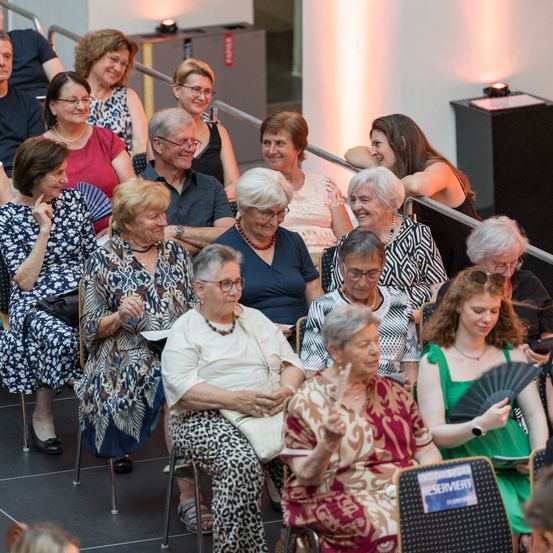 Eine Gruppe älterer Frauen sitzt in Stühlen, lächelt und unterhält sich in einem Theater, einige halten Fächer. Der Sitzbereich ist mit Metallgeländern gesäumt, und eine Frau mit Brille hält einen Fächer.