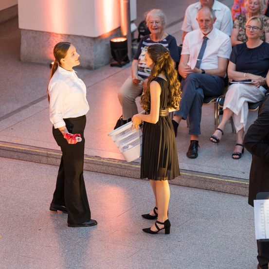 Zwei Frauen stehen sich in einem Raum gegenüber. Eine hält einen Korb. Hinter ihnen sitzt ein Publikum älterer Menschen in Stühlen und schaut zu.