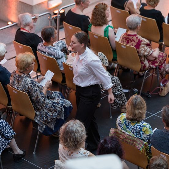 Eine Indoor-Veranstaltung, bei der eine Gruppe älterer Frauen auf Stühlen sitzen und einer Frau vor ihnen zuhören, die spricht und aus einem Blatt Papier liest.