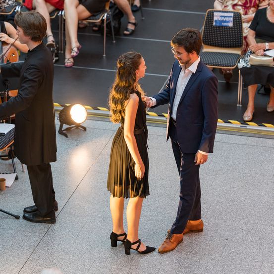 Ein Mann im Anzug spricht mit einer Frau in einem schwarzen Kleid auf der Bühne, mit einem Cellisten im Hintergrund.