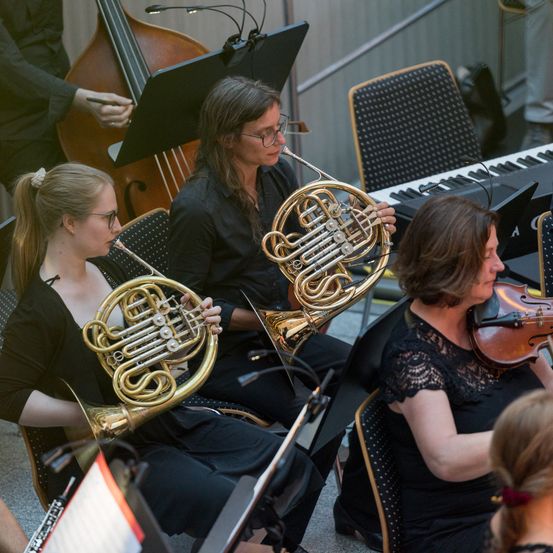 Eine Gruppe von Musikern, darunter eine Geigerin, eine Cellistin und zwei Hornisten, spielt. Sie sitzen in einem Raum, mit einem Klavier im Hintergrund.
