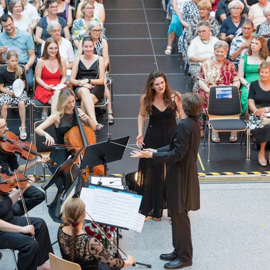 Ein klassisches Musikensemble spielt auf der Bühne, mit Geigern und einem Cellisten. Eine Frau in einem schwarzen Kleid steht neben dem Dirigenten und führt ein Gespräch. Das Publikum, das im Hintergrund sitzt, beobachtet aufmerksam die Aufführung.