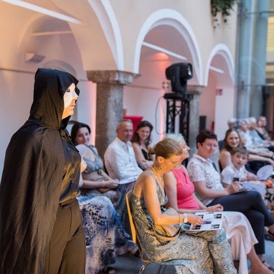 Eine Person in einem schwarzen Umhang mit einer weißen Maske steht vor einem sitzenden Publikum. Einige Publikumsmitglieder halten Papiere, während andere Getränke haben. Die Szene ist gut beleuchtet.