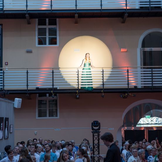 Eine Frau steht auf einem Balkon, beleuchtet von einem Scheinwerfer. Unten beobachtet sie ein großes Publikum, fasziniert. Der Ort ist ein Gebäude mit großen Fenstern und einem Bogeneingang.