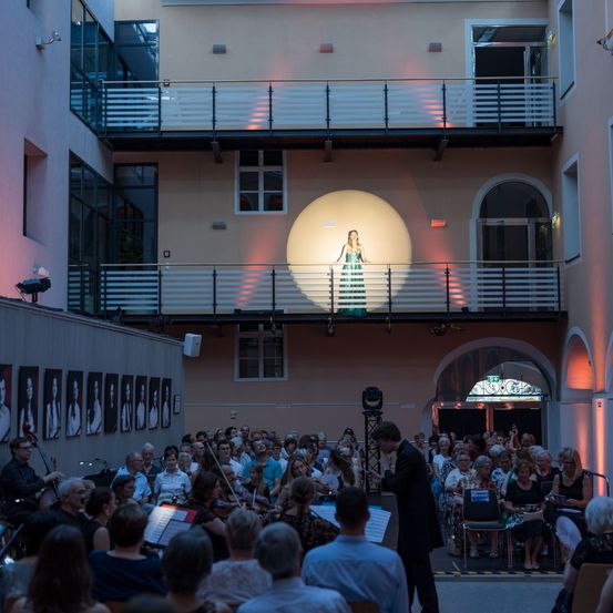 Ein klassisches Musikkonzert mit einem Orchester im Innenhof. Eine Solistin steht auf einem Balkon, beleuchtet von einem Scheinwerfer. Das Publikum sitzt vor dem Orchester.