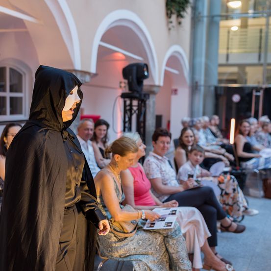 Ein Künstler in einem schwarzen Umhang und einer Maske steht vor einem Publikum, das auf Stühlen sitzt, während eine Frau vorne ein Buch in der Hand hält. Die Umgebung zeichnet sich durch gewölbte Türen und einen Hintergrund eines Gebäudes aus.