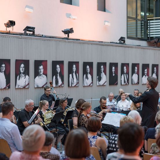 Ein Symphonieorchester spielt vor einem Publikum mit gerahmten Porträts von Musikern an der Wand.