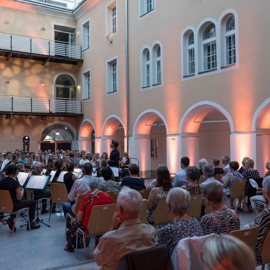Eine große Gruppe von Menschen versammelt sich in einem Innenauditorium. Einige sitzen auf Stühlen, andere stehen. Es gibt eine Gruppe von Menschen, die Musikinstrumente spielen, und eine Person steht vor ihnen. Der Raum ist mit warmer Beleuchtung erhellt.