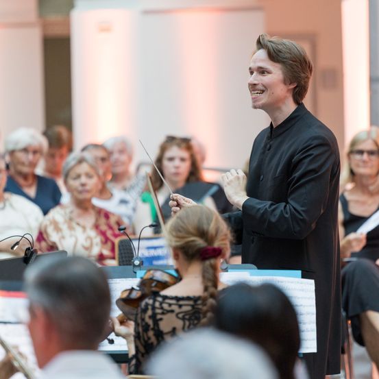 Ein männlicher Dirigent steht vor einem Orchester, lächelt, hält einen Taktstock und dirigiert die Musiker. Ein Publikum sitzt hinter dem Orchester und beobachtet.