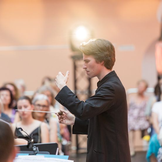 Ein Mann steht vor einem Orchester, hält einen Taktstock und dirigiert. Das Publikum sitzt im Hintergrund, einige tragen Brillen. Ein Scheinwerfer beleuchtet den Dirigenten.